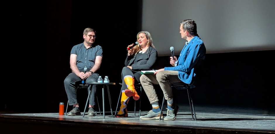 Three speakers with microphones in spotlight on stage before an empty cinema screen.