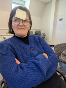 A woman in a navy sweater with a post-it noted saying 'Euchre' on her forehead.