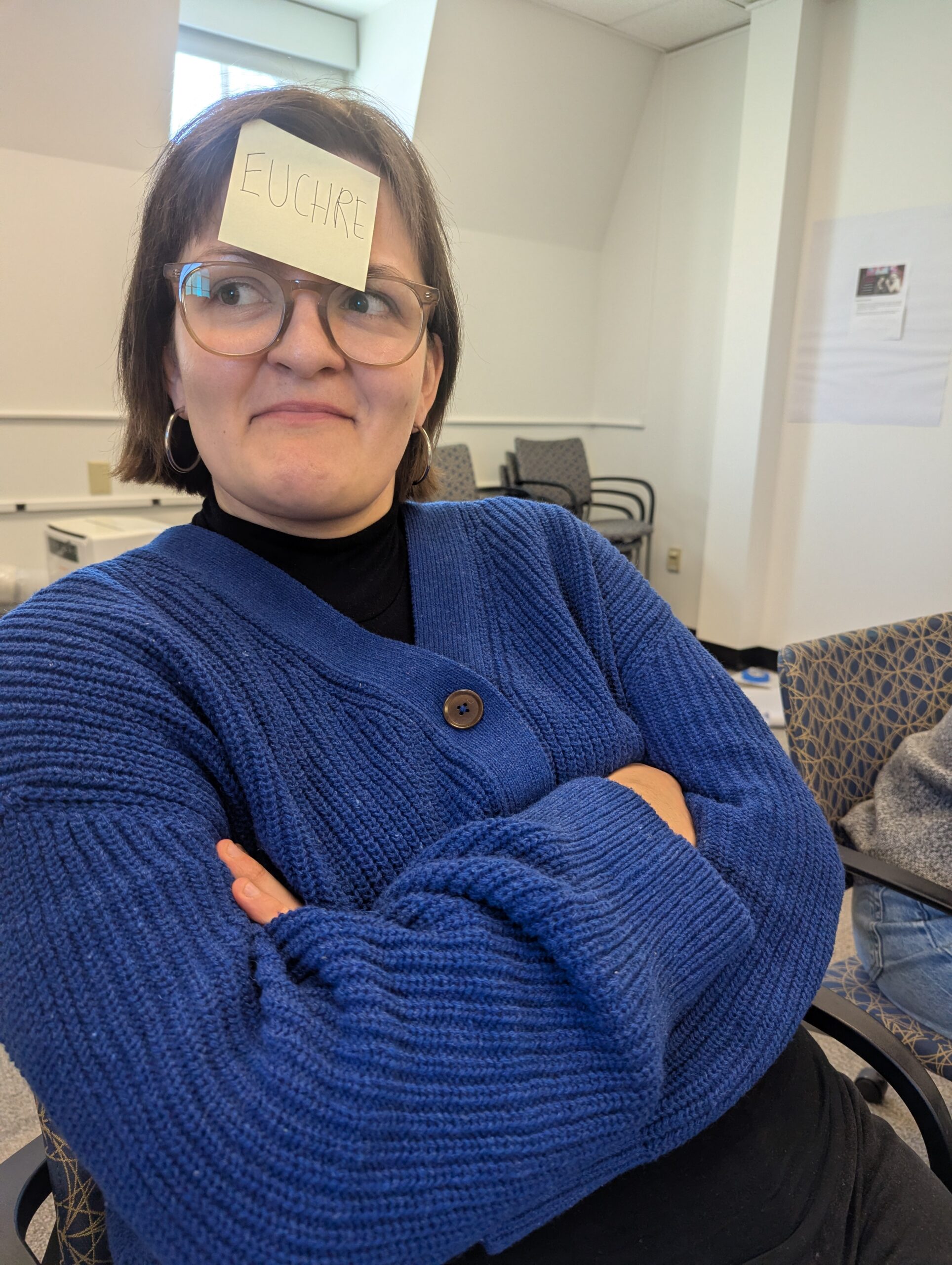 A woman in a navy sweater with a post-it noted saying 'Euchre' on her forehead.