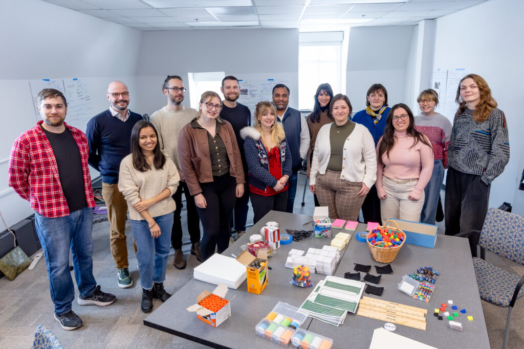 Group photo of 14 workshop participants arranged in two rows behind a table for of game design supplies.